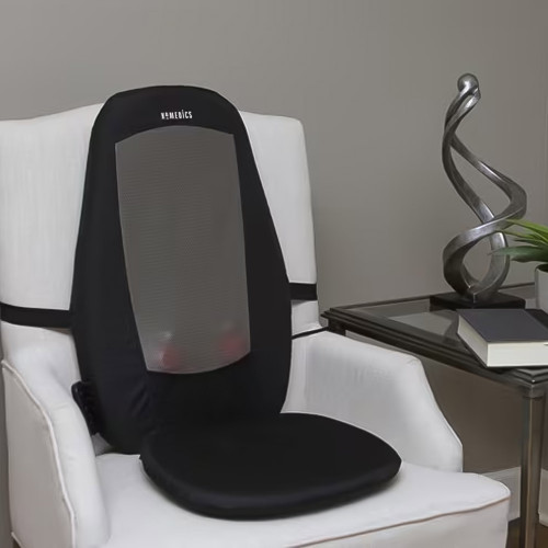 A black Shiatsu Massage Cushion with a grey mesh back panel, strapped onto a white armchair in a modern living room setting.