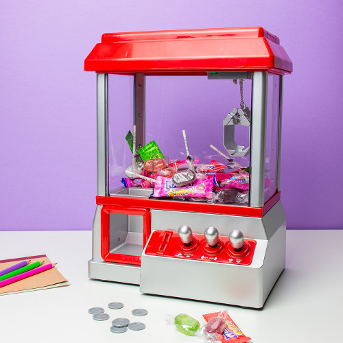 The red and silver Candy Grabber, filled with sweets, on a white desk next to play coins and coloured pencils.