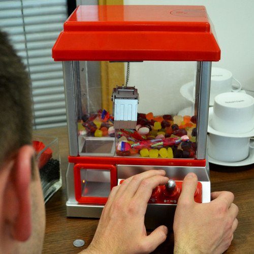 A person uses the joystick on a red Candy Grabber machine, filled with colourful sweets and sat on an office desk.