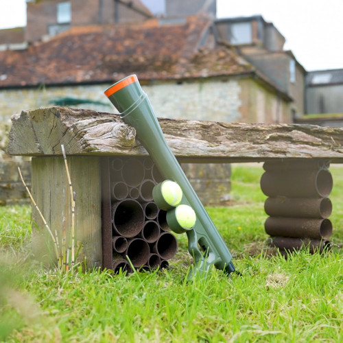 The green Dog Ball Launcher, holding two tennis balls, leans against a rustic wooden bench in a country garden.