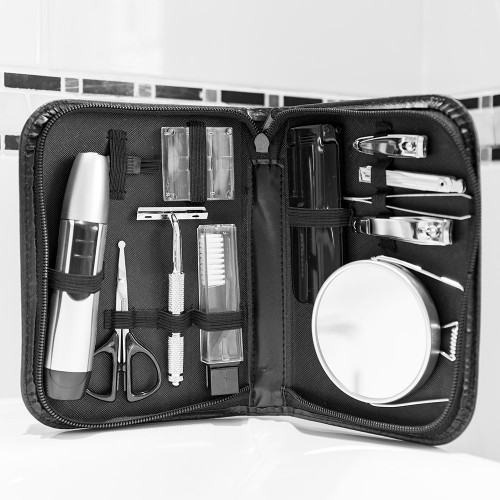 An open 10-piece grooming kit in its black carry case, resting against a tiled bathroom wall. Photographed in black and white.