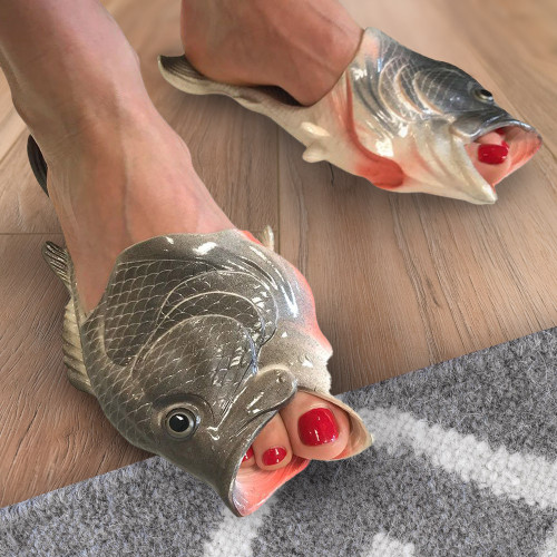 A person wears Fishy Slippers shaped like fish, the open mouths wrapped around their red-painted toes; they stand on a wooden floor next to a gray rug.