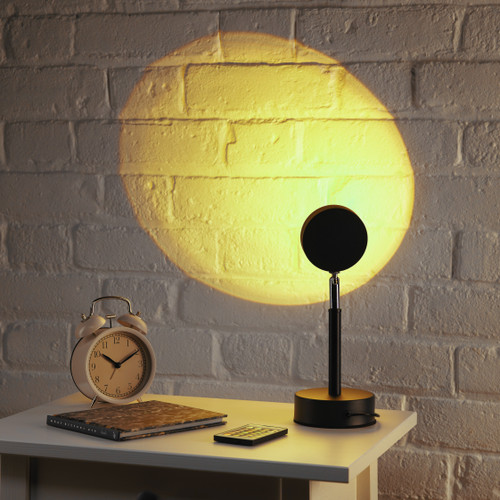 The Sundown Mood Lamp on a bedside table casts a warm yellow sunset glow onto a white brick wall next to an alarm clock.
