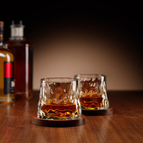 Two whiskey glasses filled with amber liquid sit on dark wood coasters on a bar top. There are bottles behind it.