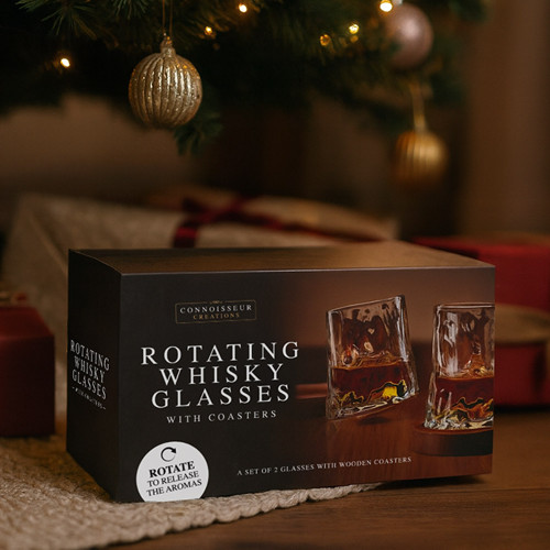 The gift box for Rotating Whisky Glasses sits on a woven mat under a decorated Christmas tree, surrounded by presents.