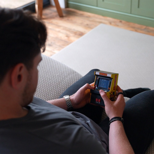 A man relaxing on a sofa while playing the yellow PAC-MAN mini arcade machine.