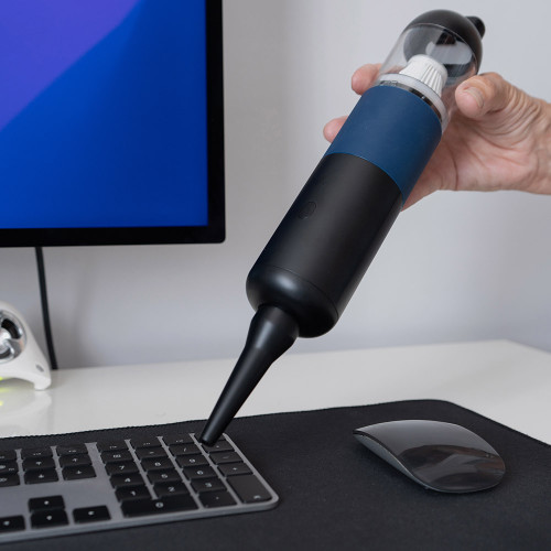 A hand holds the portable mini vacuum cleaner, cleaning between the keys of a computer keyboard on a white desk.