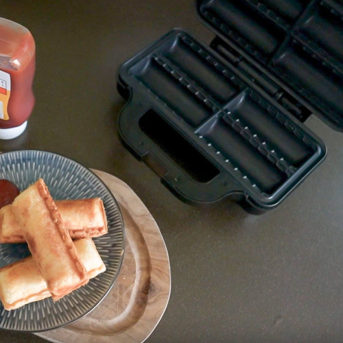 The open Sausage Roll Maker on a kitchen worktop next to a plate of freshly cooked golden-brown sausage rolls with ketchup.