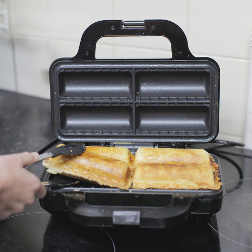 Someone uses tongs to lift a golden-brown sausage roll from the open Sausage Roll Maker.