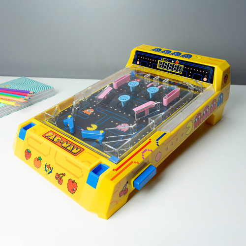A bright yellow tabletop Pac-Man pinball machine on a white desk beside coloured pencils and notebooks.