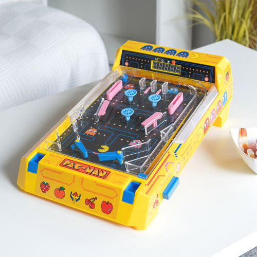 A yellow Pac Man Pinball machine on a white table next to a bowl of sweets, with a bed and a plant in the background.