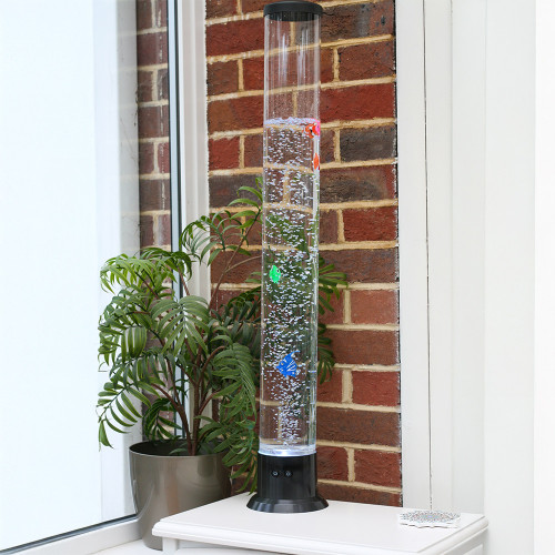 The Bubble Fish Tower, filled with water and toy fish, bubbling on a white table next to a houseplant and a brick wall.