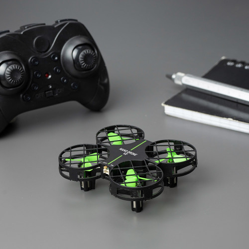 A small black-and-green Nano Drone V2 quadcopter sits on a gray surface next to a black game controller, a black notebook, and a pen.