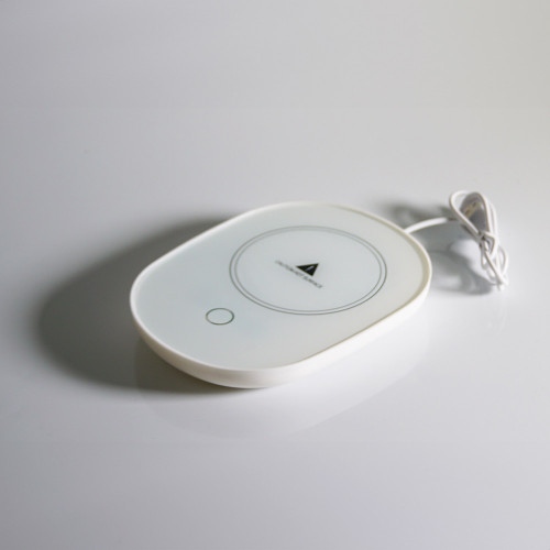 The white, oval InGenious USB Cup Warmer on a plain grey surface, showing its circular heating plate and touch power button.