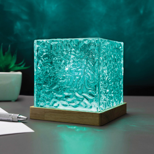 The Northern Lights Projector, a textured glass cube on a wood base, sits on a desk. It casts a turquoise glow on the wall.