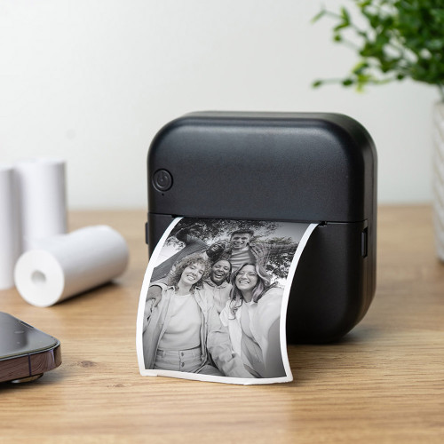 A compact black Thermal Phone Printer sits on a wooden desk, printing a black and white selfie of a group of smiling friends.