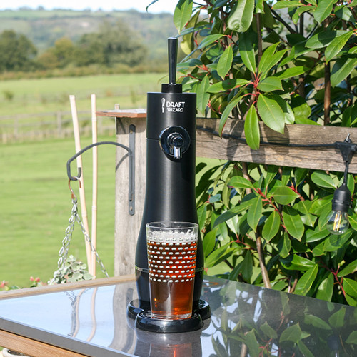 A Draft Wizard beer dispenser on a garden patio table with a fresh pint of beer, overlooking the sunny countryside.