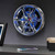The Neon Rim Wall Clock, styled as a chrome car wheel with spanner hands and blue lights, hanging on a modern grey wall.