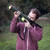 A man in a maroon hoodie aims the green Dog Ball Launcher outdoors, shouldering it like a bazooka with tennis balls loaded.