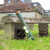 The green Dog Ball Launcher, holding two tennis balls, leans against a rustic wooden bench in a country garden.