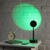 The Sundown Mood Lamp sits on a white bedside table next to an alarm clock, projecting a green circle of light on the wall.