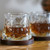 A whiskey glass filled with an amber drink, sitting on a dark wooden coaster. A second glass is behind it.