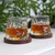 Two whiskey glasses with an amber drink sit on dark wood coasters on a white table, with a leafy plant in the background.