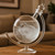 The Globe Storm Glass sits on a wooden table, with white, cloudy crystals swirling inside the clear liquid.