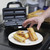 A hand takes a golden-brown sausage roll from a plate. The open Sausage Roll Maker is behind it.
