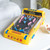 A yellow Pac Man Pinball machine on a white table next to a bowl of sweets, with a bed and a plant in the background.