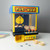 The Pac-Man arcade claw crane on a white desk. A Pac-Man claw hangs over prize balls, with toy coins and an open capsule nearby.