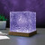 The textured, cube-shaped Northern Lights Projector sits on a wooden base, casting a vibrant purple glow on a desk.