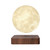 A realistic, glowing moon lamp with a textured surface levitates over a square, dark wood-grain base.