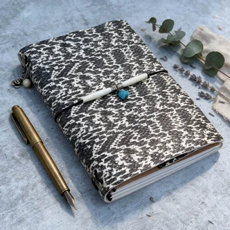 Black and white snakeskin-patterned leather journal on a writing desk with a brass pen, eucalyptus, and dried lavender.