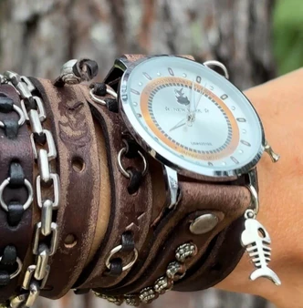 Distressed dark brown leather wrap watch worn on a wrist with layered straps, metal rings, silver-tone case, and fishbone charm.