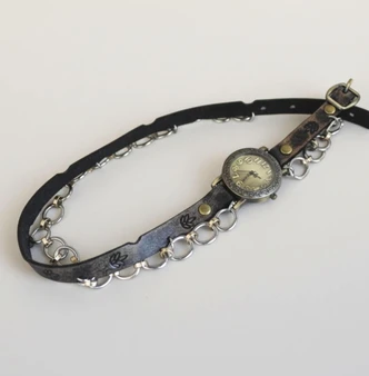 Black distressed full-grain leather women's watch with silver chain detail and decorative leaf stamps, laid flat on white background