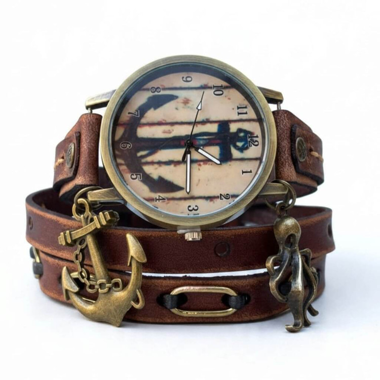 Close-up of vintage-style anchor watch face surrounded by distressed brown leather, decorative brass-tone metal loops, and nautical charms