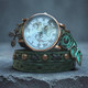 Styled close-up of women's green leather wrap watch with world map watch face, copper-tone cross charm, and verdigris leaf clock charm on a blue-gray background.