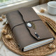 Taupe gray refillable leather journal with crystal-style and leaf charms on a woven placemat beside a cup and brass pen.