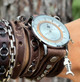 Distressed dark brown leather wrap watch worn on a wrist with layered straps, metal rings, silver-tone case, and fishbone charm.