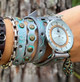 Metallic silver gray leather wrap watch worn on a wrist with layered straps, antique brass studs, turquoise details, and decorative teardrop charm.