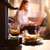 Caffe Borbone Didi Machine For Espresso Paper Pods, coffee machine brewing a fresh espresso shot with a glass cup, alongside a plate of cookies, in a cozy setting with a woman working on a laptop in the background.