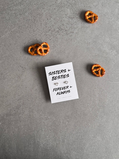 Sterling silver heart knot earrings on a 'Sisters + Besties' message card. The earrings are shown next to some pretzels.