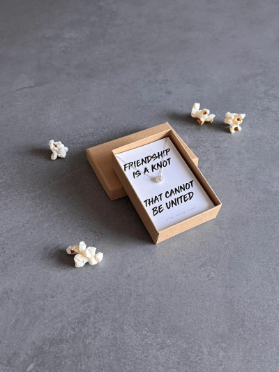 Sterling silver friendship knot necklace on a white message card and in a Kraft gift box. The necklace is shown surrouded by popcorn. Sterling silver friendship knot necklace on a white message card and in a Kraft gift box. The necklace is shown surrouded by popcorn.