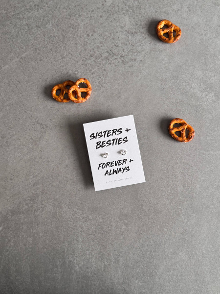 Sterling silver heart knot earrings on a 'Sisters + Besties' message card. The earrings are shown next to some pretzels.