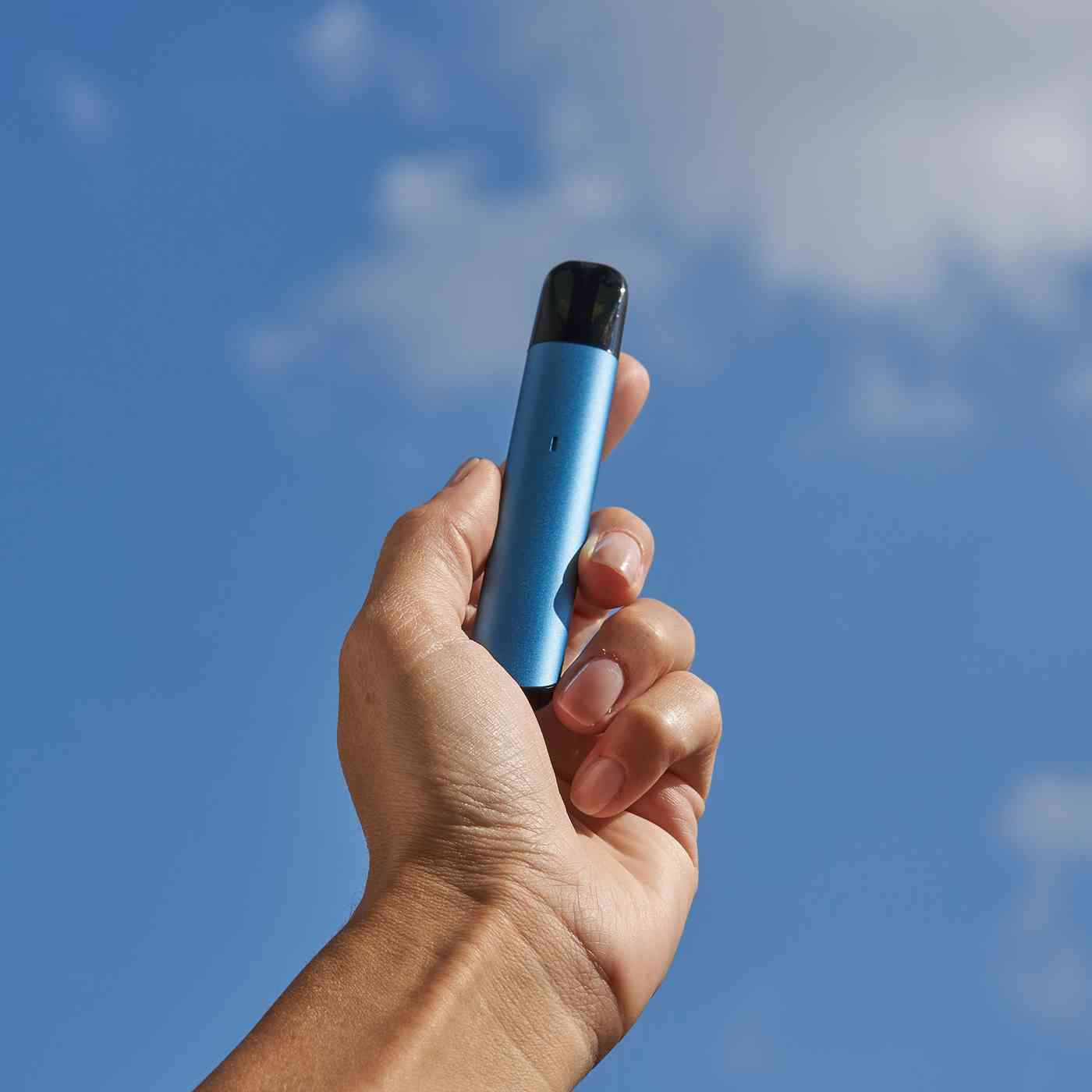 A hand holding a slim blue MTL pod kit against a blue sky, the type of efficient device that uses less e-liquid per day under the new vaping duty.