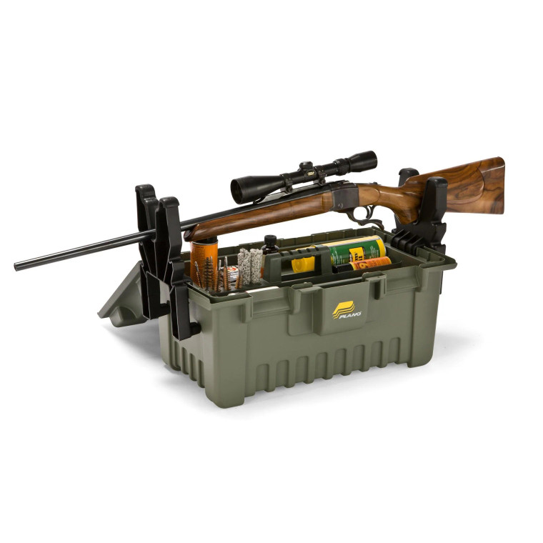 Plano green hunting storage and cleaning box with an integrated gun rest, shown holding a scoped rifle and various cleaning supplies including brushes, oils, and tools. The sturdy plastic case features built-in clamps to secure the firearm for maintenance.