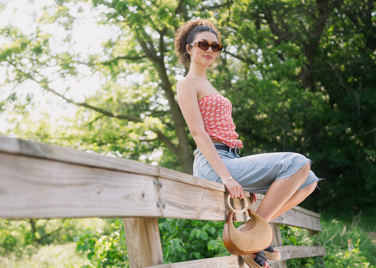 Person sitting on a wooden fence outdoors, holding a woven handbag, with trees in the background.