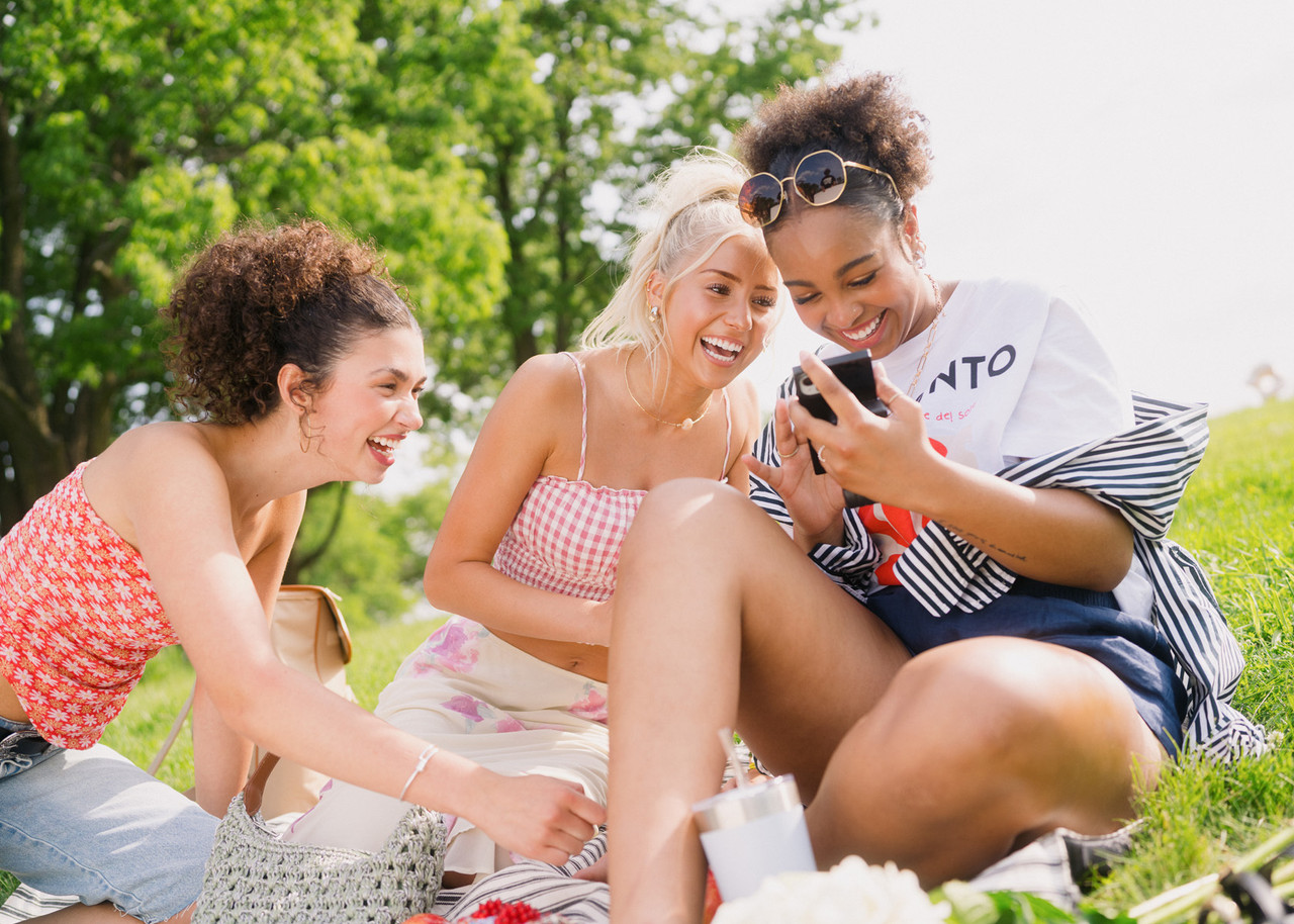 Three people sitting on a blanket on a grassy hill, looking at a smartphone together outdoors.