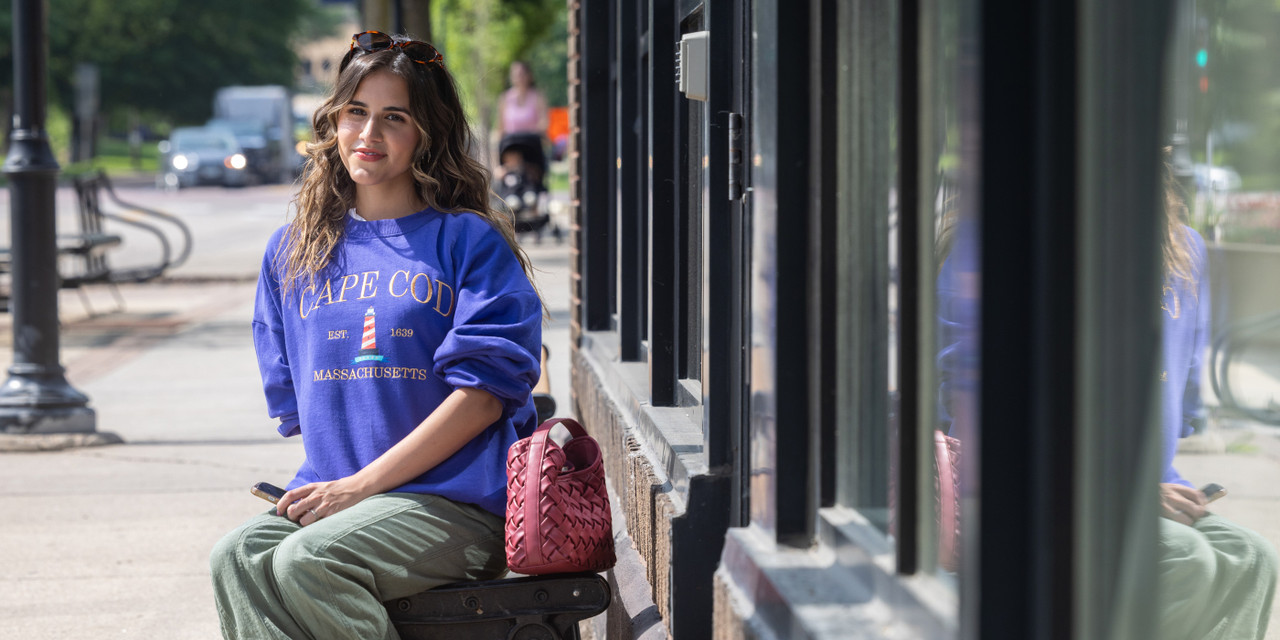 Person sitting on a bench along a sidewalk, wearing a blue sweatshirt, with a handbag beside them and storefront windows nearby.